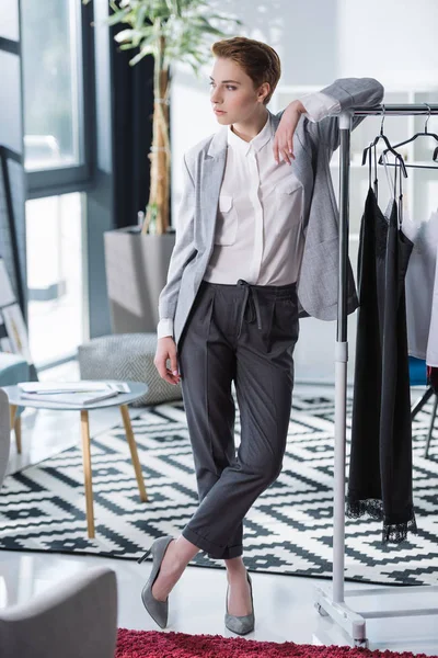 Beautiful young fashion designer leaning on bar with dresses and looking away — Stock Photo