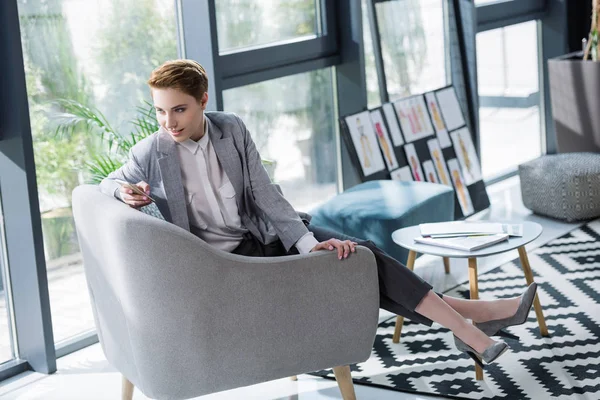 Attraktive junge Modedesignerin mit Smartphone im Sessel im Büro — Stockfoto