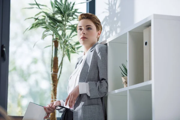 Attractive young businesswoman with book leaning back on bookshelves at office — Stock Photo