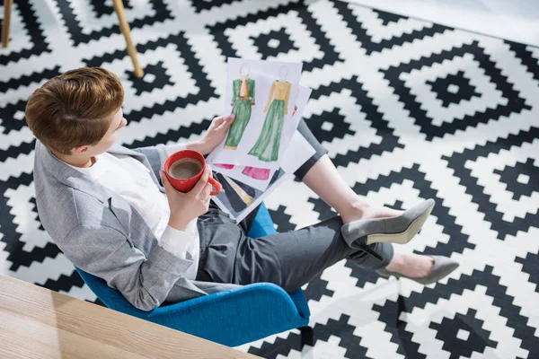 High angle view of young fashion designer with cup of coffee looking at sketches — Stock Photo