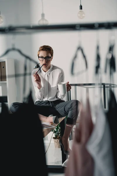 Stylish fashion designer with cup of coffee sitting on desk and looking at dresses — Stock Photo