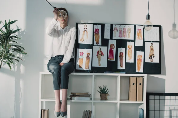 Stylish fashion designer sitting on bookshelves and looking at light bulb — Stock Photo