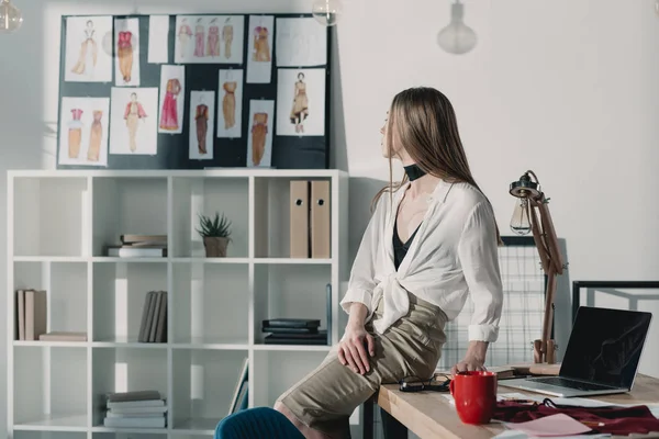 Stylish fashion designer sitting on table at office and looking at sketches — Stock Photo