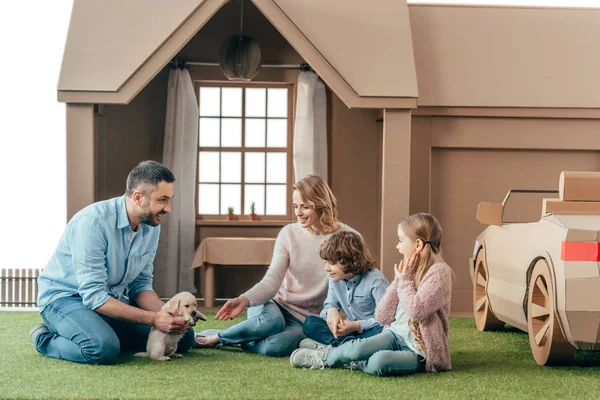 Schöne junge Familie sitzt mit ihrem Welpen auf dem Hof eines Papphauses — Stockfoto