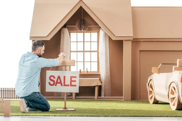 Чоловік вішає вивіску продажу перед картонним будинком ізольовано на білому — стокове фото