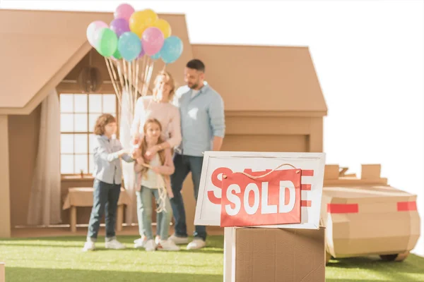 Letrero vendido con la familia joven en el patio de su nueva casa de cardbord borrosa en el fondo aislado en blanco - foto de stock