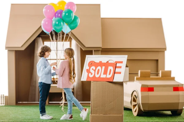 Kid presenting balloons to girlfriend in front of cardboard house with sold signboard on foreground isolated on white — Stock Photo
