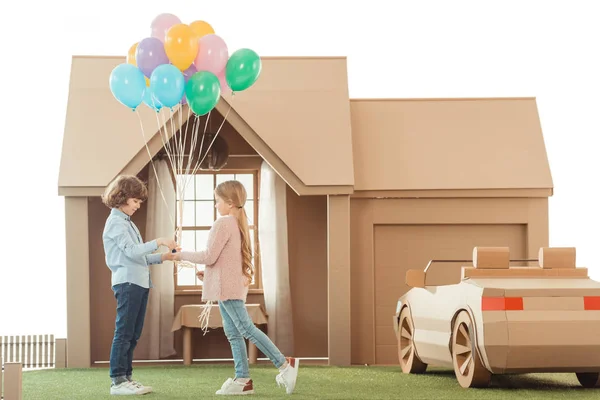 Kid presenting balloons to girlfriend in front of cardboard house isolated on white — Stock Photo