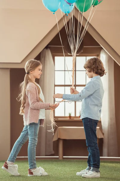 Kid presenting balloons to beautiful little girlfriend in front of cardboard house — Stock Photo