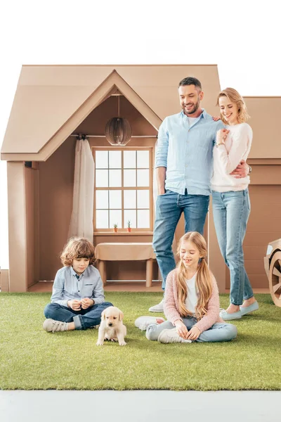 Young family with cute puppy on yard of cardboard house isolated on white — Stock Photo