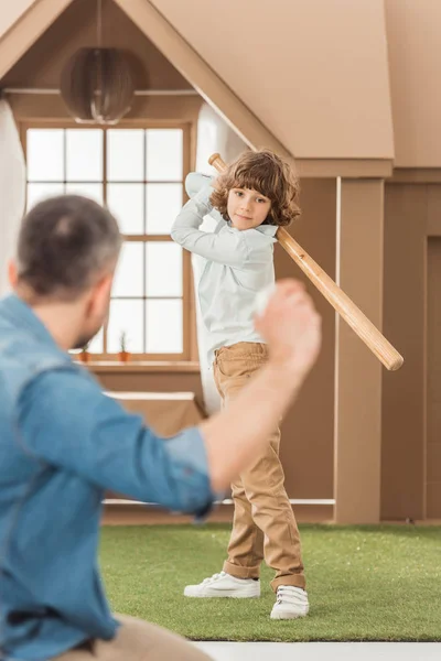 Отец учит своего маленького сына играть в бейсбол перед картонным домиком — стоковое фото