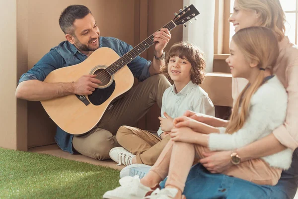 Счастливый красивый отец играет на гитаре для детей и жены в новом картонном доме — стоковое фото