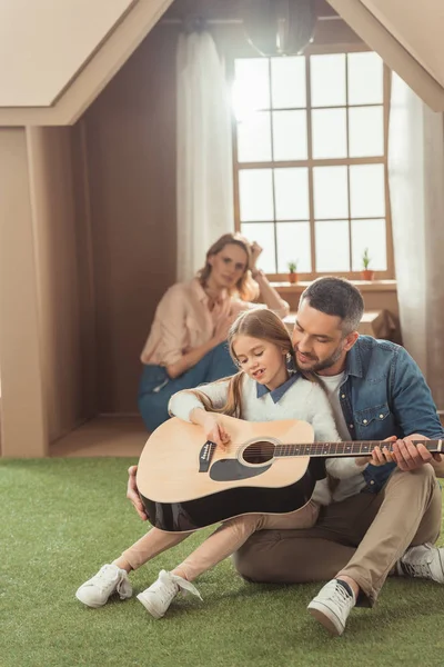 Красивий батько і дочка грають на гітарі на траві перед картонним будинком з матір'ю, що сидить всередині — стокове фото
