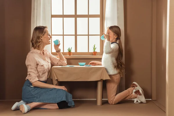 Laughing mother and daughter having tea party in cardboard house — Stock Photo