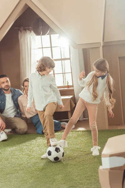 Geschwister spielen Fußball auf Hof eines Papphauses, während Eltern sie anschauen — Stockfoto