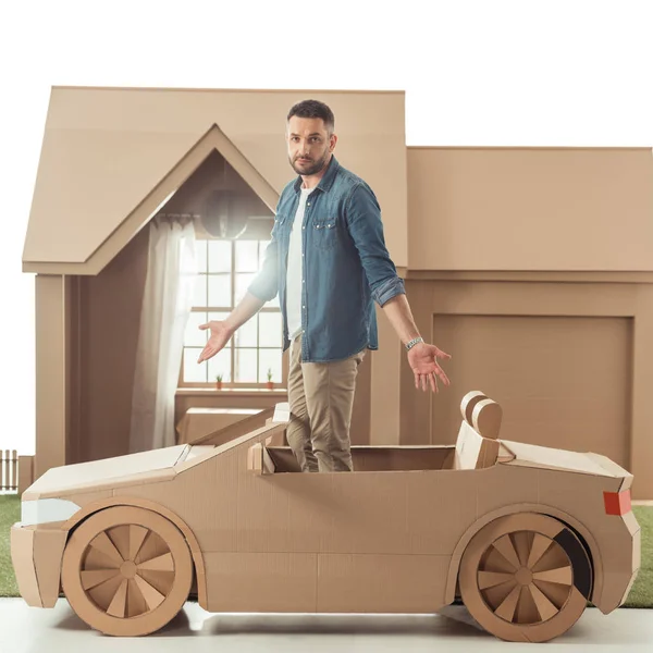 Bel homme dans la voiture en carton devant la maison en carton isolé sur blanc — Photo de stock