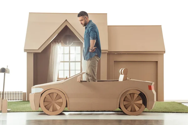 Vue latérale de l'homme en voiture en carton devant la maison en carton isolé sur blanc — Photo de stock