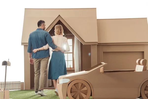 Вид сзади пары, обнимающейся перед картонным домом, изолированным на белом — стоковое фото