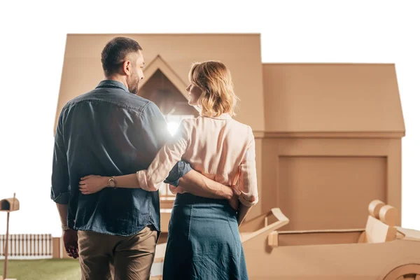 Вид сзади счастливой пары, обнимающейся перед картонным домом, изолированным на белом — стоковое фото