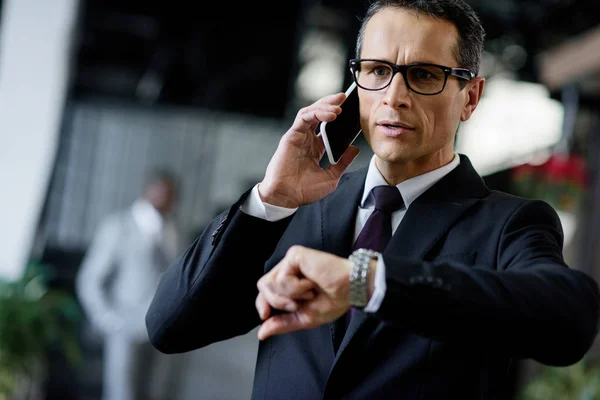 Retrato de homem de negócios verificando o tempo enquanto fala no smartphone — Fotografia de Stock