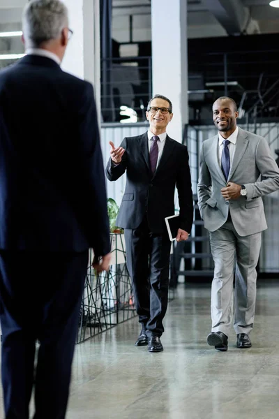 Multiethnisch lächelnde Geschäftsleute in Anzügen gehen im Büro spazieren — Stockfoto