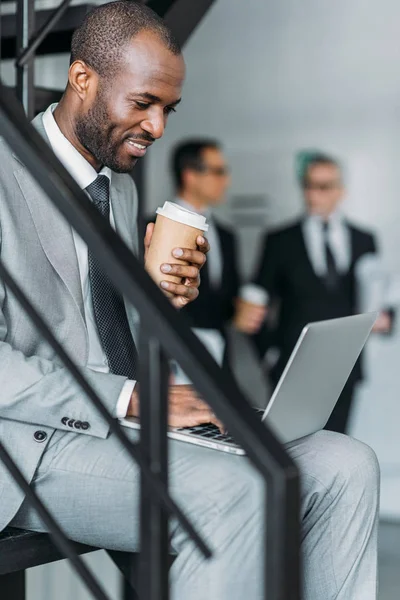 Selective focus of smiling african american with coffee to go businessman working on laptop — Stock Photo