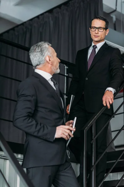 Бізнесмени в костюмах мають розмову, йдучи вниз по сходах в офісі — стокове фото