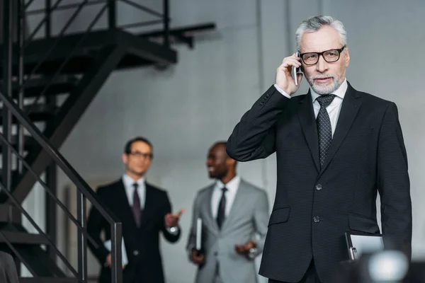 Enfoque selectivo del hombre de negocios hablando en el teléfono inteligente con sus colegas detrás en la oficina - foto de stock