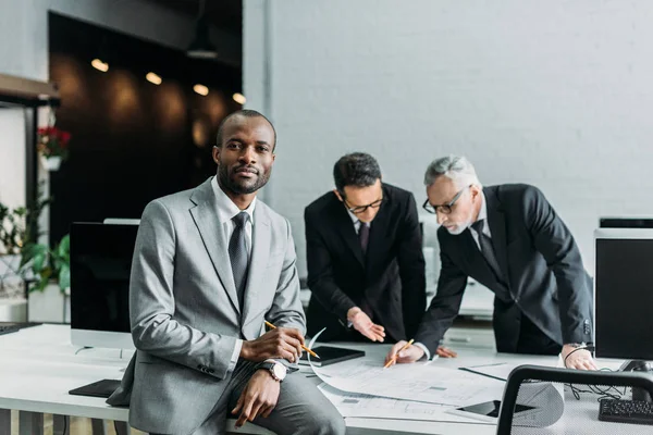 Afrikanischer amerikanischer Geschäftsmann blickt in die Kamera, während Geschäftskollegen über Arbeit im Büro diskutieren — Stockfoto