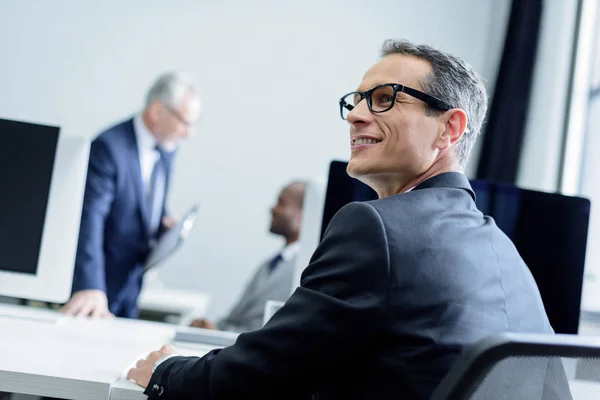 Selektiver Fokus eines lächelnden Geschäftsmannes mit Brille, der im Büro wegschaut — Stockfoto