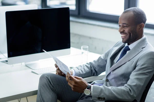 Sorridente uomo d'affari africano americano che legge il giornale sul posto di lavoro — Stock Photo