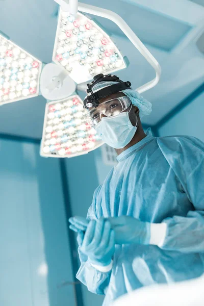 Vista inferior del cirujano afroamericano con guantes médicos y mirando hacia otro lado - foto de stock