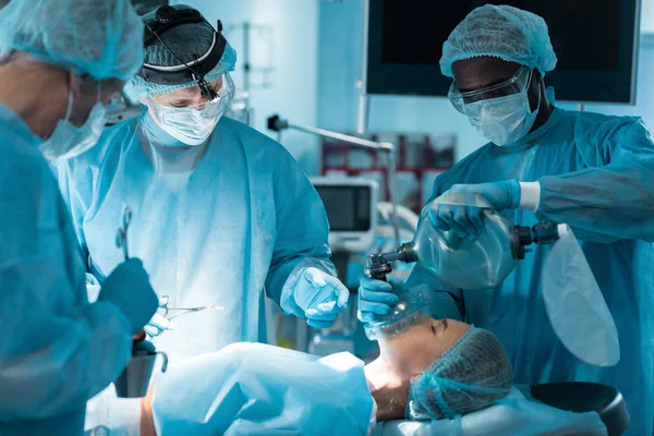 Anestesista afro-americano segurando máscara de oxigênio acima do paciente na sala de cirurgia — Fotografia de Stock