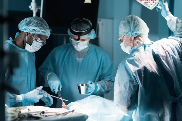 Cirujanos multiétnicos en máscaras médicas operando al paciente en quirófano - foto de stock