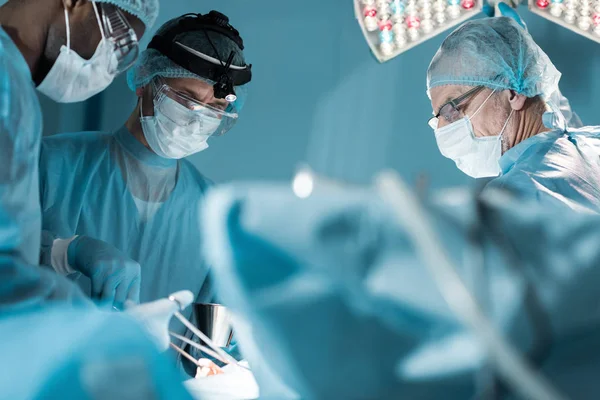 Cirujanos multiculturales en máscaras médicas operando al paciente en quirófano - foto de stock