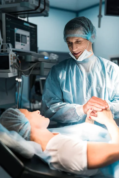 Médico sonriente y paciente tomados de la mano en la sala de cirugía - foto de stock