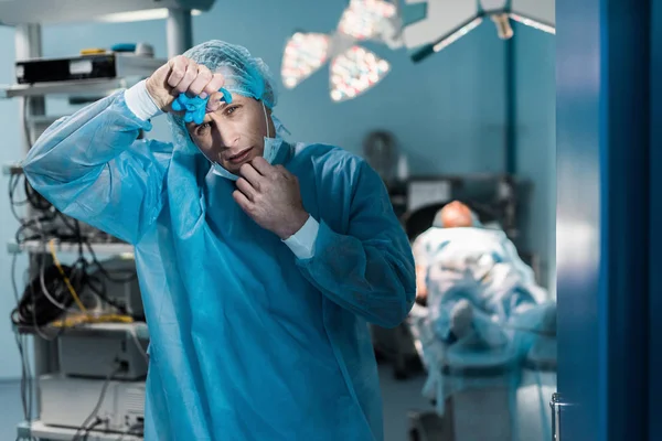 Tired doctor removing medical mask and touching forehead in surgery room — Stock Photo