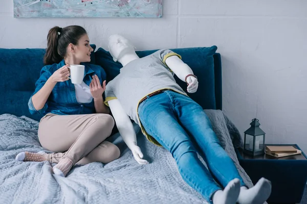 Young woman with cup of coffee talking to mannequin while resting on bed, unrequited love concept — Stock Photo