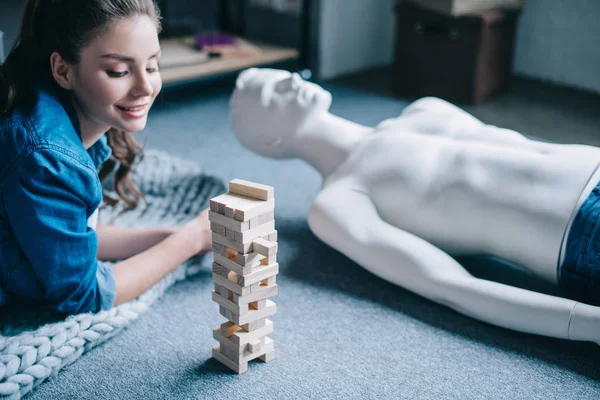 Beautiful woman lying near mannequin and blocks wood game on floor at home, loneliness concept — Stock Photo