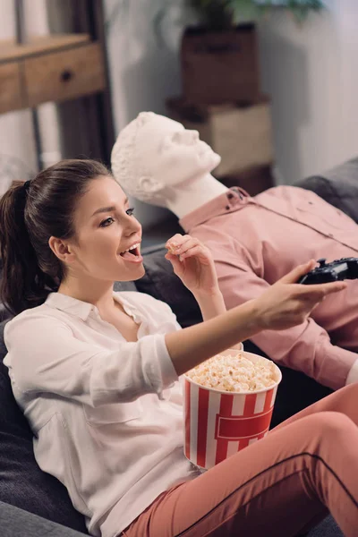Side view of woman eating popcorn while playing video game with manikin near by, loneliness concept — Stock Photo