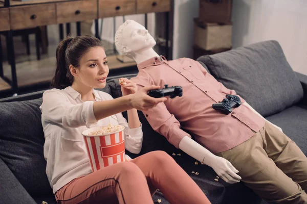 Jeune femme manger du pop-corn tout en jouant à un jeu vidéo avec mannequin à proximité à la maison, concept de solitude — Photo de stock