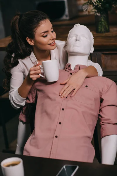 Junge Frau mit Tasse Kaffee umarmt Schaufensterpuppe zu Hause, perfektes Beziehungstraum-Konzept — Stockfoto