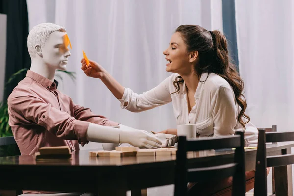 Joven mujer jugando juego junto con laico muñeca en la mesa con tazas de café, concepto de soledad - foto de stock