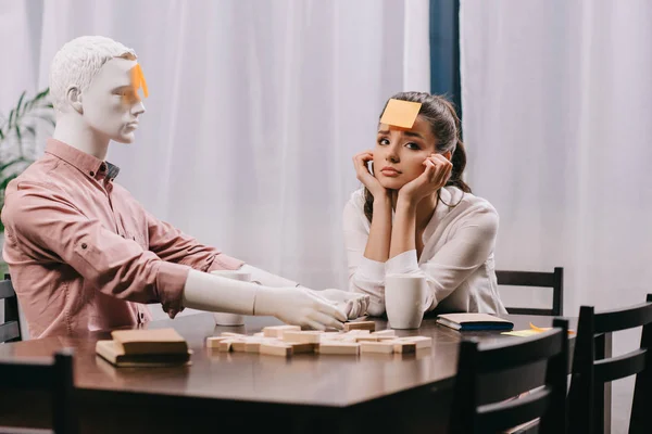 Sad young woman with sticky note on forehead sitting at table with mannequin, loneliness concept — Stock Photo