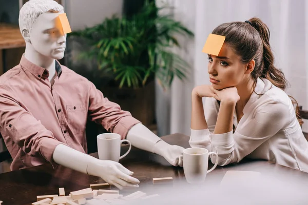 Sad young woman with sticky note on forehead sitting at table with mannequin, loneliness concept — Stock Photo