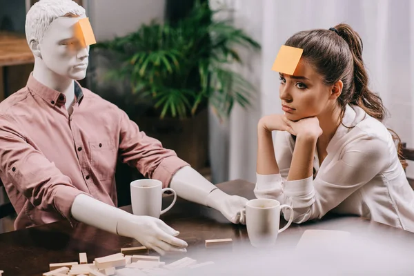 Triste joven con nota adhesiva en la frente sentado a la mesa con maniquí, concepto de soledad - foto de stock