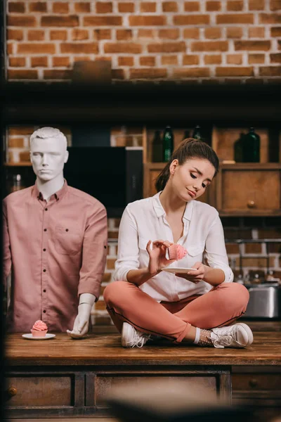 Woman with cupcake sitting on table with mannequin behind, loneliness concept — Stock Photo