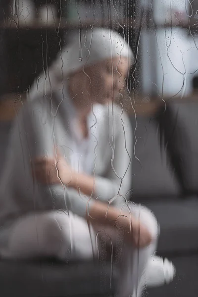 Close-up view of glass with raindrops and sick mature woman behind — Stock Photo