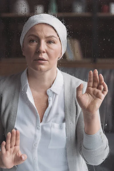 Kranke reife Frau im Halstuch blickt durch ein Glas mit Regentropfen in die Kamera — Stockfoto