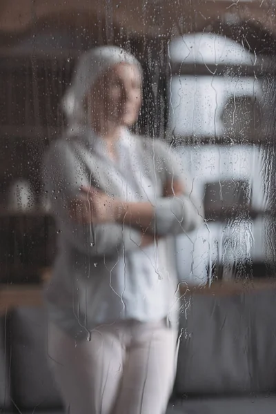 Vue rapprochée du verre avec des gouttes de pluie et femme mûre malade debout derrière — Photo de stock